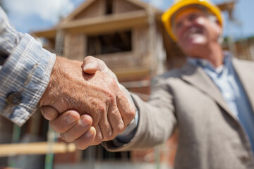 Close-up of a construction professional and a businessman or client shaking hands on a residential building site, symbolizing agreement and partnership in real estate development.