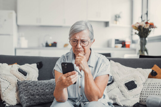 Elderly woman sits on a sofa at home looking at her phone with disappointment. She receives bad news while shopping online, feeling frustrated and cautious about digital transactions.