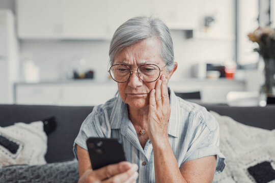 Elderly woman sits on a sofa at home looking at her phone with disappointment. She receives bad news while shopping online, feeling frustrated and cautious about digital transactions.