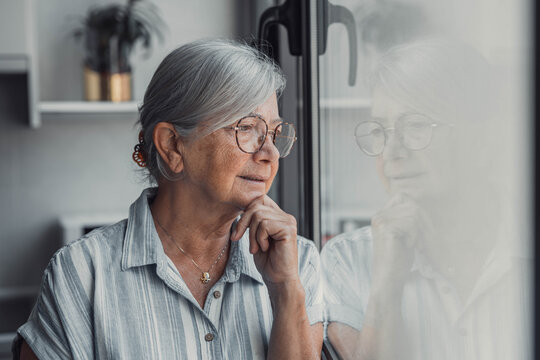 Elderly woman looks out the window with a worried expression. She reflects on her thoughts and feels concerned about her day while standing at home.