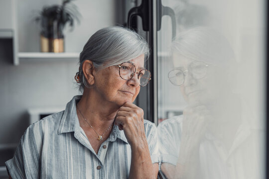 Elderly woman looks out the window with a worried expression. She reflects on her thoughts and feels concerned about her day while standing at home.
