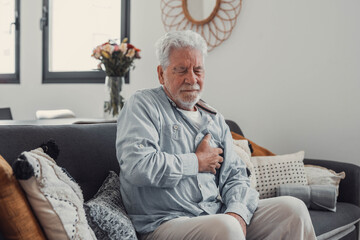 Old man sits on a sofa clutching his chest in distress. He appears anxious and fatigued while...