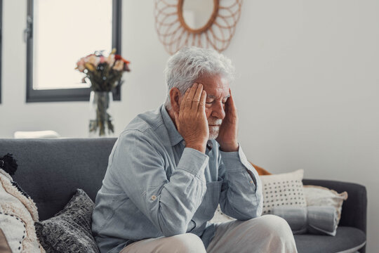 Mature man sits on a sofa holding his head in discomfort. He experiences headache and fatigue while coping with stress and illness at home.