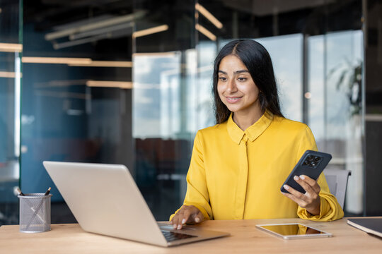A young Indian businesswoman is working in the office at a laptop and holding a mobile phone in her hand