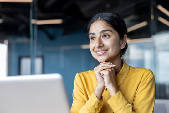 Close-up photo of a young Indian businesswoman in a yellow shirt sitting at a desk in the office, looking forward thoughtfully and smiling