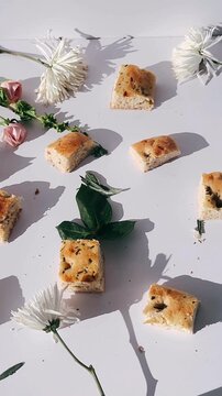 soft herb focaccia bread pieces scattered across a white surface with fresh flowers, leaves, crumbs, and gentle natural shadows creating a calm, rustic food scene.