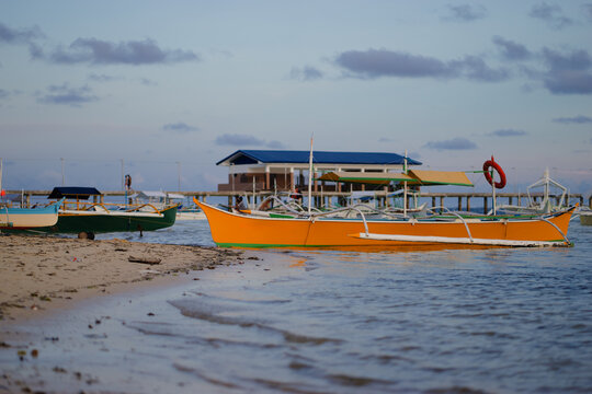 Brightly painted wooden boats rest along a sandy beach with a pier and building nearby, under a serene evening sky. - Powered by Adobe