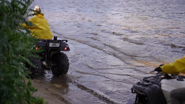 Slow motion. Lake shore rider in yellow jacket cruising shallow water, lone quad rolling from muddy bank toward gentle waves, overcast sky and green trees framing quiet
