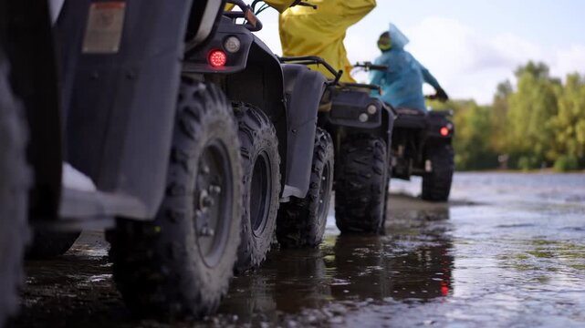 Slow motion. Closeup of ATV wheels splashing water, yellow and blue jacketed riders, low angle shot highlights muddy tires, wet trail reflections and forest backdrop,
