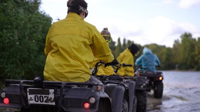 Rear view ATV riders by river, three riders in yellow and blue rain jackets riding muddy shore, peaceful forest backdrop, convoy teamwork and guide led