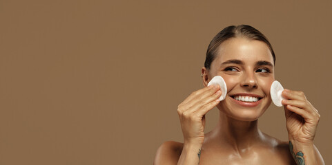 Smiling woman cleansing face with cotton pads on brown background