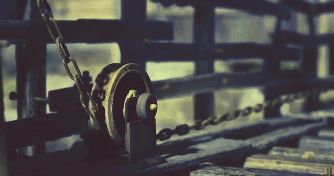 A rusty pulley hangs from a weathered wooden structure, connected by a chain. The abandoned setting shows signs of decay and age, creating a rustic atmosphere.