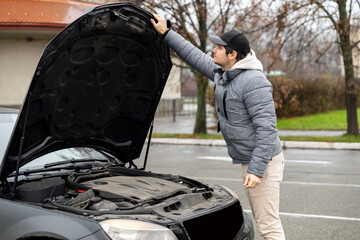Young man inspecting car engine under open hood in parking lot