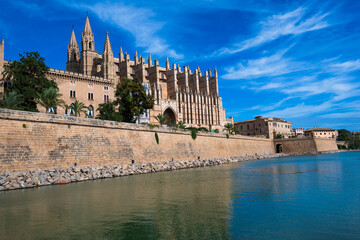 Waterfront cityscape with historic buildings, palm trees and a modern sculpture rising from the water under a clear blue sky