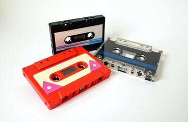 Obraz premium Three vintage audio cassettes displayed on a clean white surface. One red cassette is prominent, with a black and a transparent cassette positioned behind it, evoking nostalgic music listening.