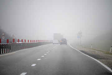 Vehicles moving along highway in thick fog with low visibility and traffic signs, illustrating dangerous driving conditions and road safety in poor weather.