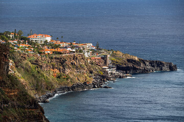 Clifftop coastal village in Madeira with orange-tiled roofs overlooking the Atlantic Ocean, rugged volcanic cliffs, and a seaside swimming platform carved into the rocks