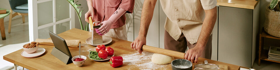 Website banner of man rolling dough while woman with prosthetic arm grating cheese in kitchen, fresh vegetables and digital tablet on wooden table