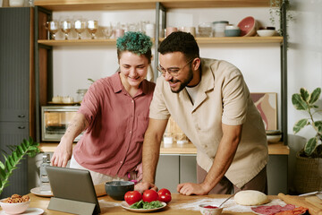 Young adult Caucasian woman and man smiling while preparing food together in kitchen, looking at tablet screen, surrounded by fresh vegetables and dough on countertop