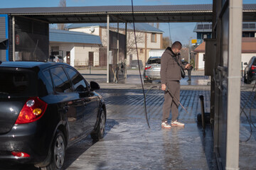 Man washes car at self-service car wash in sunny afternoon with clear sky and modern buildings in...
