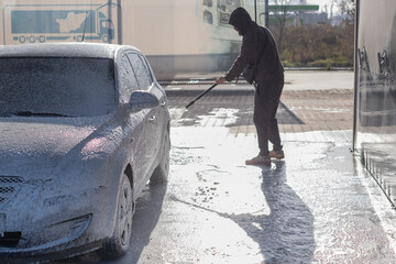 Man washes car with foam at a self-service car wash in bright sunlight during the day near a parking lot