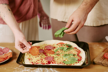 Unrecognizable woman and man preparing homemade pizza together, arranging pepperoni slices and fresh spinach leaves on uncooked dough in kitchen