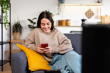 Young happy teenage girl using mobile phone while relaxing on sofa at home while watching TV