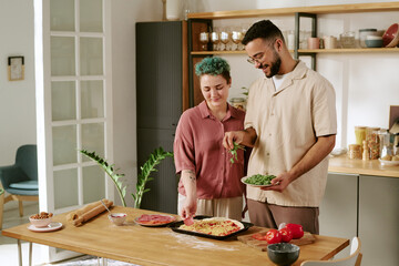 Young adult Caucasian woman and man preparing homemade pizza together in modern kitchen, smiling and interacting while adding fresh ingredients to baking tray