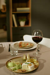 Cheese platter with assorted cheeses, walnuts, pear slices and honey served on plate next to glass of red wine and water glass on table, pizza slice in background