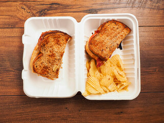 A sandwich is sitting in a plastic container on a wooden table. The sandwich is made with two slices of bread and a piece of meat. The container is open, revealing the sandwich inside.