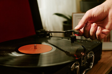 Male hand adjusting tonearm on vinyl record player, spinning black vinyl disc on turntable, indoor setting with blurred background and houseplant visible