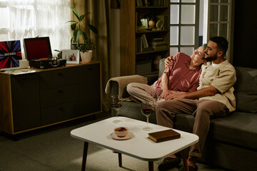 Middle aged Caucasian woman and middle aged man sitting on sofa embracing and smiling, relaxing together at home with two glasses of red wine on table in foreground