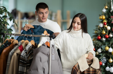 Guy helps a young woman with the choice of a winter upper one time. Couple standing in a shop decorated for Christmas and buying warm clothes