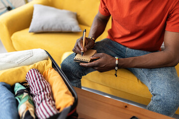 C,lose up view of young African man sitting on sofa preparing suitcase for travel, making checklist at home. Travel and vacation concept