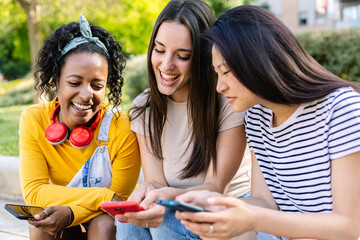 Three young multiracial women friends using mobile phone sitting outdoors - Female friendship concept with millennial people sharing social media content on smart phone device