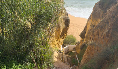 Sandy beach passage between jagged, gray rock cliffs with green grass on top, bright daylight,...