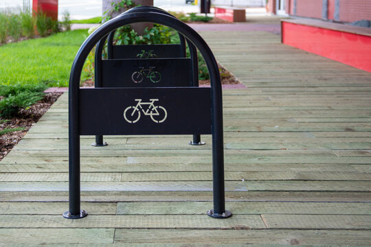 A black metal bicycle parking rack. The stationary fixture with bike cutout symbols on the arms and a curved freestanding rack is secured to a wooden deck at a park. The outdoor bike rack is empty.