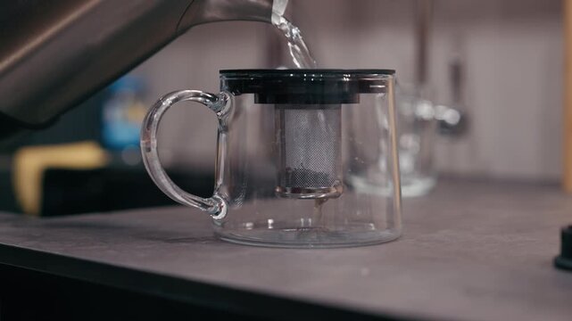Clear glass teapot with infuser on countertop, hot water is poured into it, revealing rich amber tea color, showcasing brewing process with dynamic camera movement
