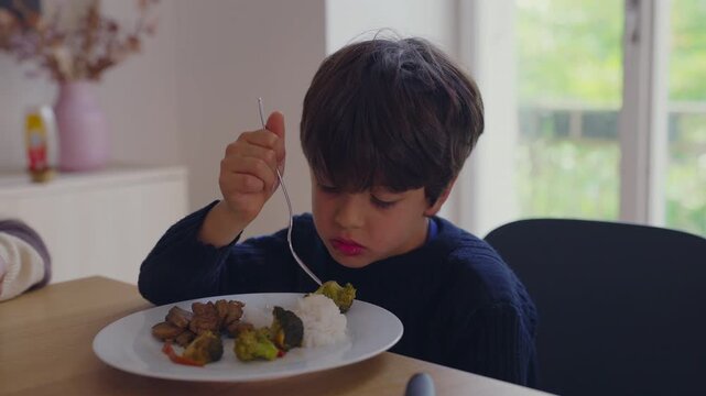 Child eating vegetables and rice at family table, focused expression and routine nourishment reflecting calm domestic life learning autonomy and everyday childhood growth,