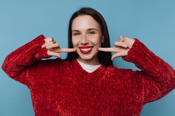 Woman smiles and poses with fingers on a blue background wearing a red sweater and bright lipstick...