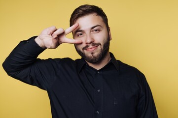 Man with beard makes peace sign in front of yellow wall during daylight while wearing black shirt and smiling at the camera