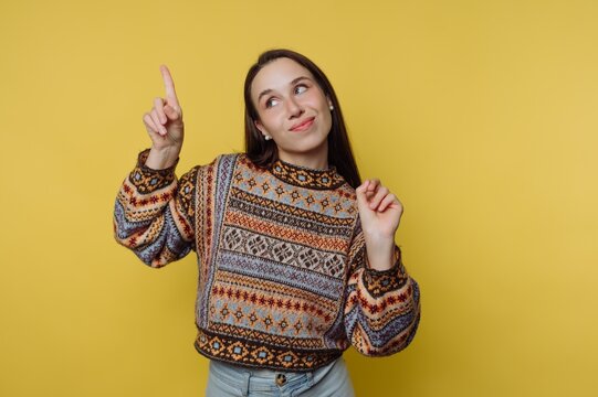 Young woman points upward with a smile against a yellow background while wearing a patterned sweater