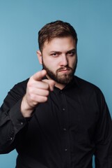 Obraz premium Man in black shirt points finger at the viewer against a light blue background while showing serious expression and focused gaze