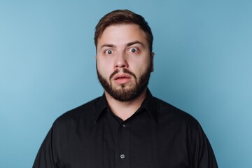 Obraz premium Man displays surprise with wide eyes and an open mouth while standing against a blue background, conveying a strong reaction