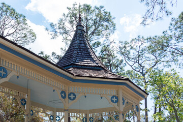 A cream colored wooden turret with blue decorative details on the wood panels. The Queen Anne style outdoor garden building with open sides and wooden posts. The roof has cedar shakes as roof tiles. 