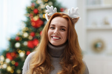 Portrait of smiling woman in reindeer headband against blurred lights at home. Merry Christmas and...