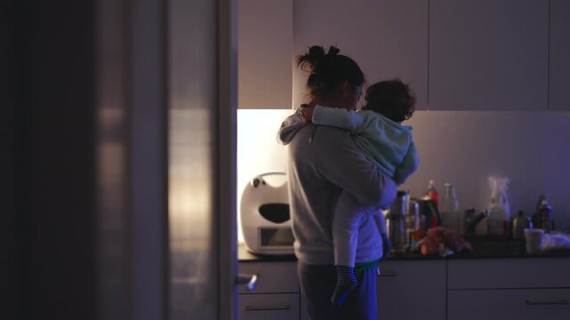 Mother holding tired toddler in dimly lit kitchen during quiet evening moment while standing near counter with household items and warm backlight creating calm indoor atmosphere