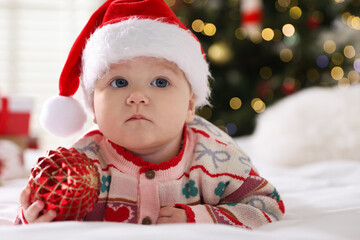 Cute little baby in Santa hat with gift box and bauble on bed at home. Christmas season
