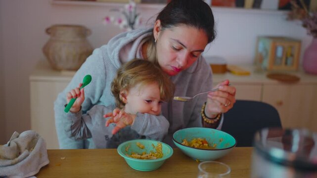 Mother offering spoon to toddler who resists feeding, gentle persistence autonomy and everyday tension revealing real caregiving dynamics during home mealtime routine