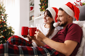Happy couple in pajamas with cups of hot drink spending time together on bed in room decorated for Christmas